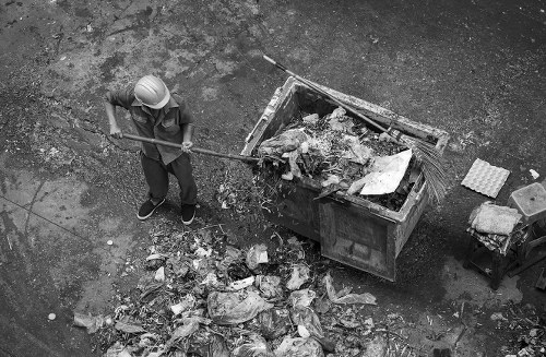 A worker wearing a safety helmet and dark clothing is using a long-handled tool to clear loose rubbish from the ground near a large industrial skip truck filled with mixed waste materials. The waste includes cardboard, plastic, and various debris, some spilling onto the asphalt surface below. The skip is positioned on a paved surface, likely an alleyway or service area in an urban setting. The scene is photographed from an elevated angle, capturing both the worker and the contents within the skip, with additional small items such as a brick and some packaging material nearby. The environment is outdoors, with a gritty, industrial atmosphere typical of rubbish removal operations in central London or similar urban areas, supporting local rubbish collection services like those offered by Office Clearance Barbican. The overall scene illustrates a typical waste clearing activity aligned with professional rubbish removal in the Barbican area, underscoring efficiency and environmental consideration in waste disposal tasks.