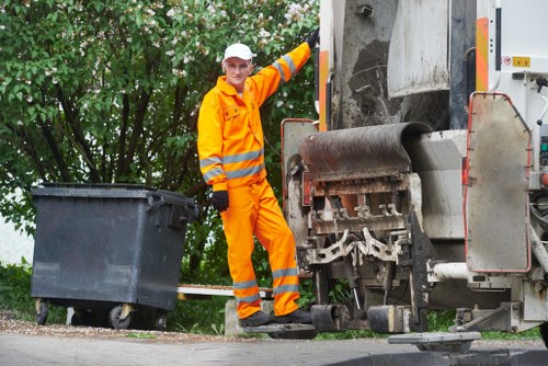 A waste collection worker dressed in high-visibility orange protective clothing, including a jacket and trousers with reflective stripes, stands beside a large commercial rubbish compactor truck on a paved street. The worker, wearing a white helmet and gloves, is smiling and appears to be in the process of overseeing or assisting with rubbish collection. The truck features a metal roller and conveyor system for compressing waste, with visible dark grime and a metallic grey finish. To the left of the worker, a black wheeled rubbish bin is positioned on the pavement, ready for collection. In the background, lush green foliage and trees are visible, indicating an outdoor urban setting, possibly in the Barbican area or nearby town. The scene suggests a professional rubbish removal operation, typical of local waste management services such as those offered by Office Clearance Barbican, carried out on a typical day of rubbish collection within a city environment.