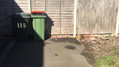 The image shows a small area of pavement adjacent to a wooden fence, with two wheelie bins positioned against the fence on the left side. One bin is dark green with the number '115' printed on it in white, and the other is also dark green with a closed lid. Both bins are made of plastic and stand upright on the concrete surface. There is a noticeable dark patch or stain on the pavement close to the bins, and the surrounding ground features a small patch of soil with sparse weeds or grass along the right edge. The wooden fence behind the bins is constructed from vertical planks, with the two sections of fencing meeting at a corner. The lighting suggests daytime, with the sunlight casting shadows from the bins onto the ground. The scene appears to be a typical rubbish collection point, consistent with services offered by Office Clearance Barbican in London.