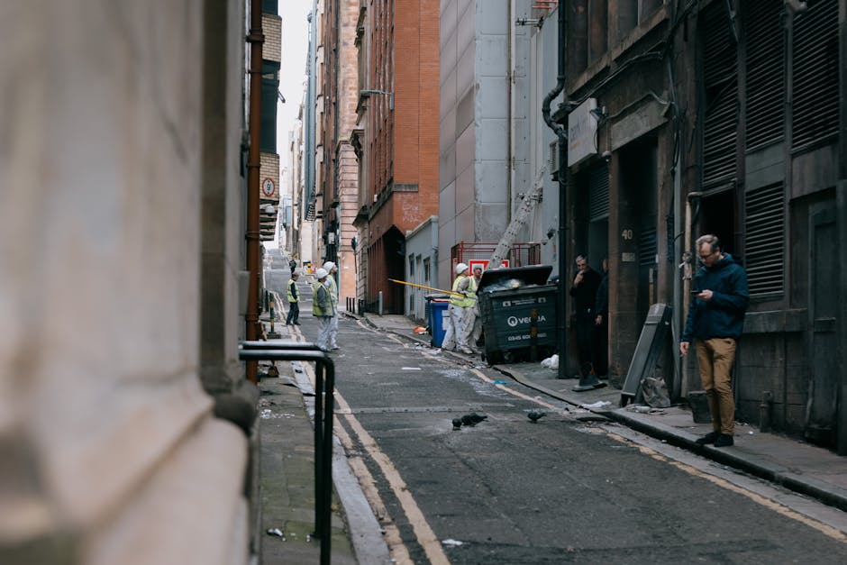 A large, weathered, beige fabric rubbish bag is positioned on the right side of a narrow, paved urban street, leaning against a textured, graffiti-marked concrete wall. The bag is filled with various