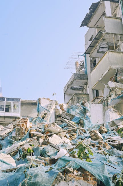 A large pile of rubble composed of broken concrete, bricks, and twisted metal rebar sits in front of a partially demolished multi-story building. The debris includes scattered bricks, shattered concre