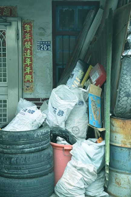 Multiple large transparent plastic bags filled with assorted waste materials, including clothing, textiles, and miscellaneous refuse, are placed on a paved sidewalk in front of a commercial establishm